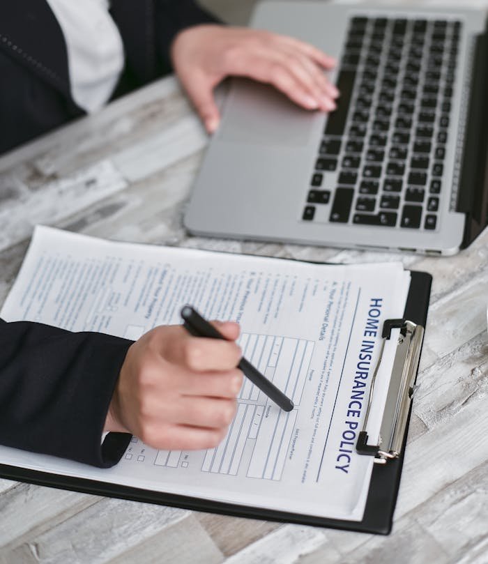 An adult reviews and signs a home insurance policy document on a clipboard next to a laptop.